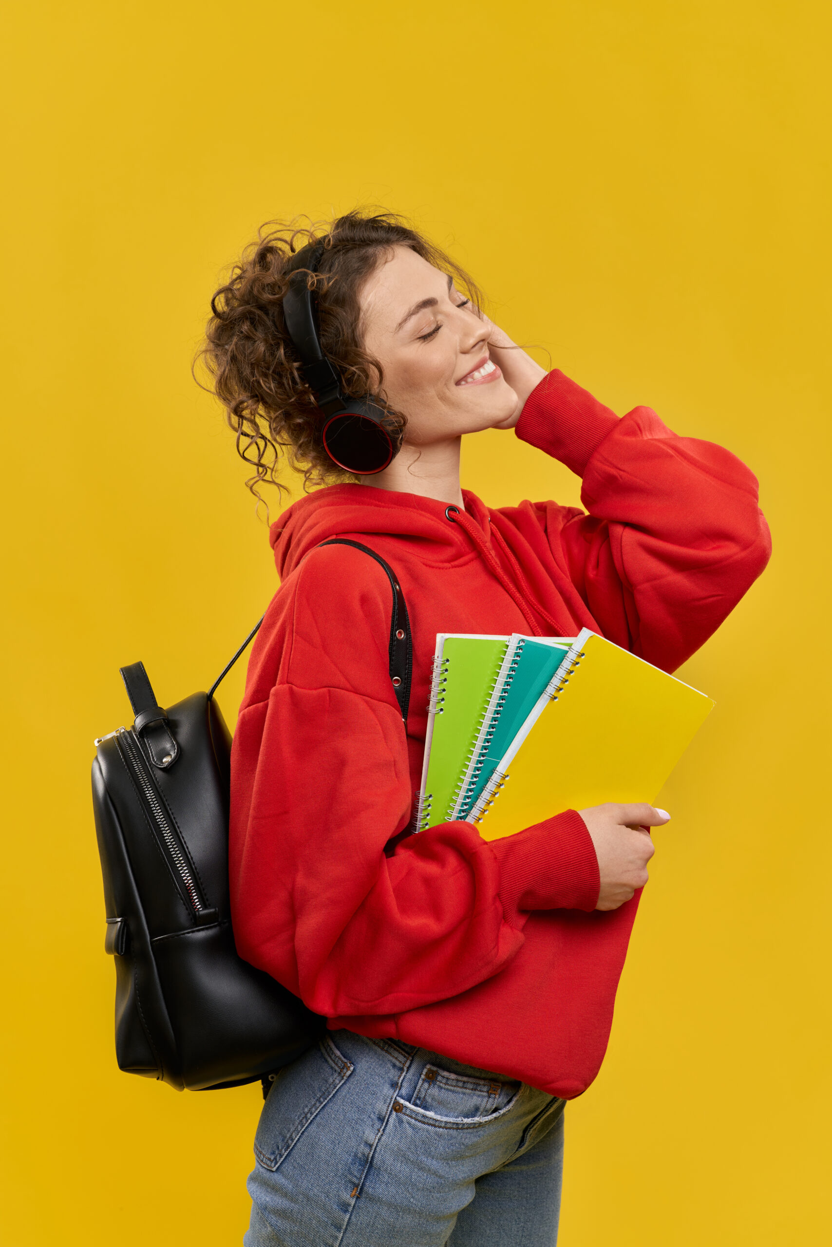 Side view of female student listening to music in earphones. Girl standing with rucksack, holding books, looking up with closed eyes, smiling. Isolated on yellow background.